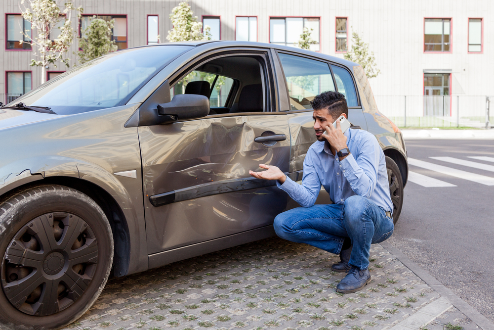 Hombre hispano viendo los daños de su vehículo y haciendo llamada para preguntar cómo reclamar a un seguro de coche